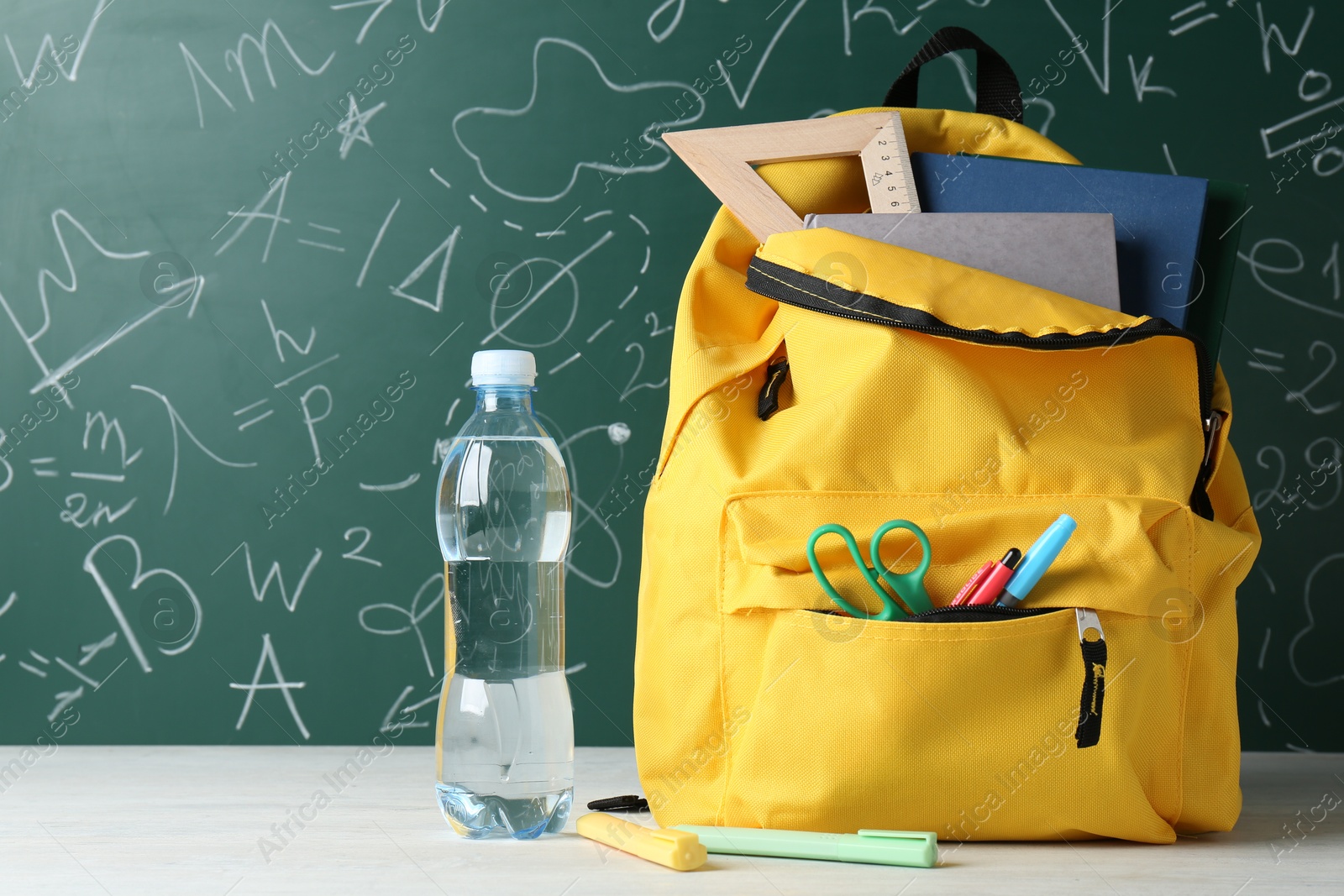 Photo of Backpack with different school stationery and bottle of water on white table against green chalkboard