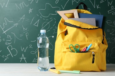 Photo of Backpack with different school stationery and bottle of water on white table against green chalkboard