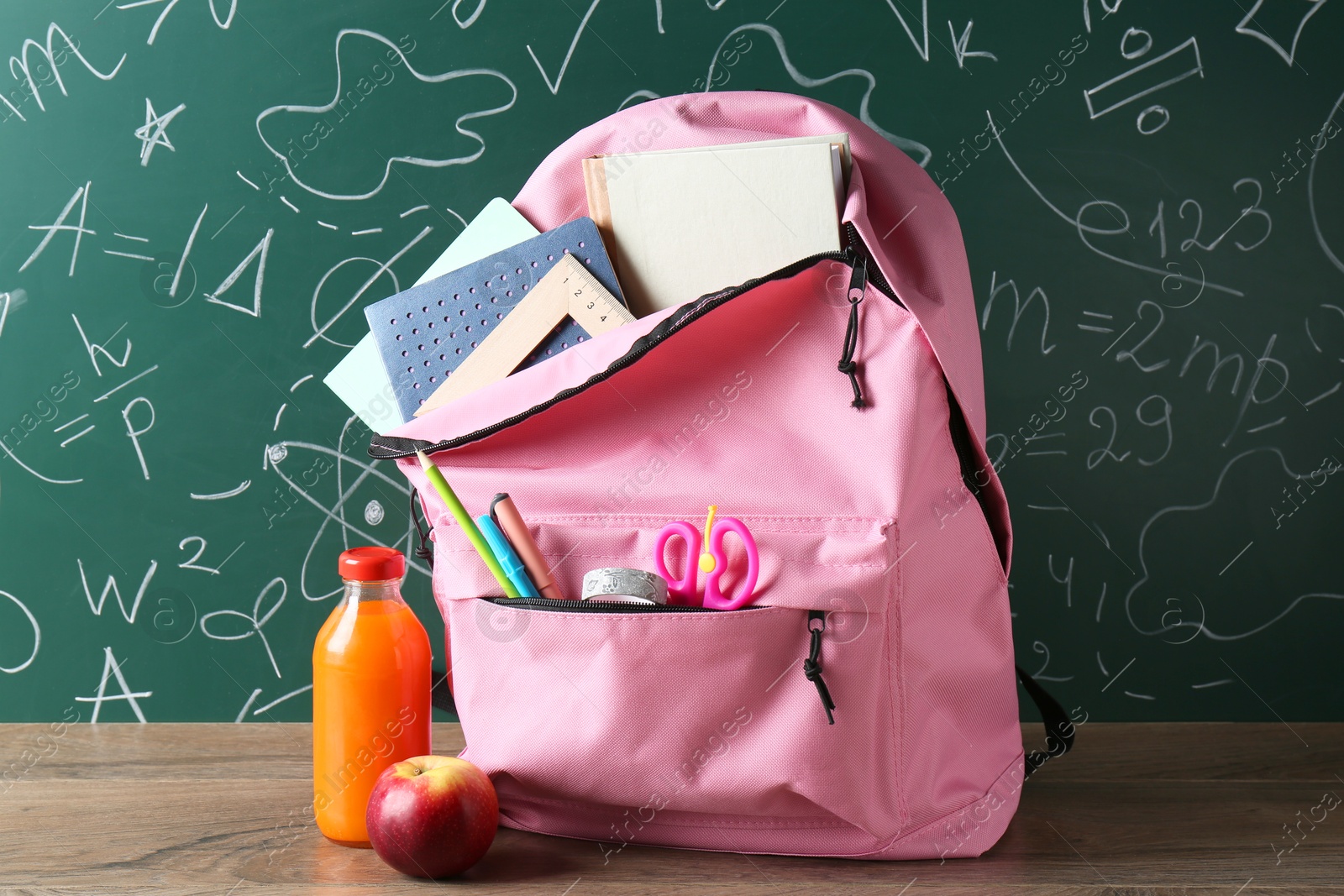 Photo of Backpack with different school stationery, apple and juice on wooden table against green chalkboard