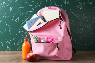 Backpack with different school stationery, apple and juice on wooden table against green chalkboard