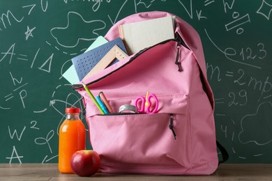 Photo of Backpack with different school stationery, apple and juice on wooden table against green chalkboard