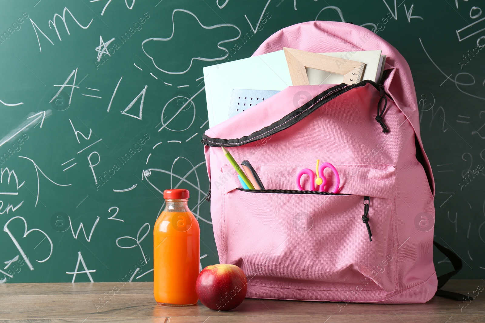 Photo of Backpack with different school stationery, apple and juice on wooden table against green chalkboard