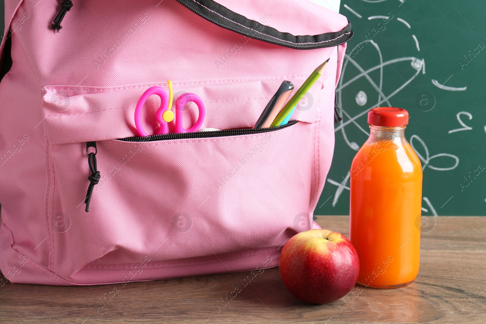 Photo of Backpack with different school stationery, apple and juice on wooden table against green chalkboard