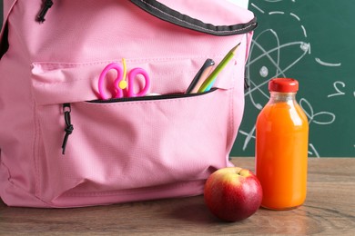 Photo of Backpack with different school stationery, apple and juice on wooden table against green chalkboard