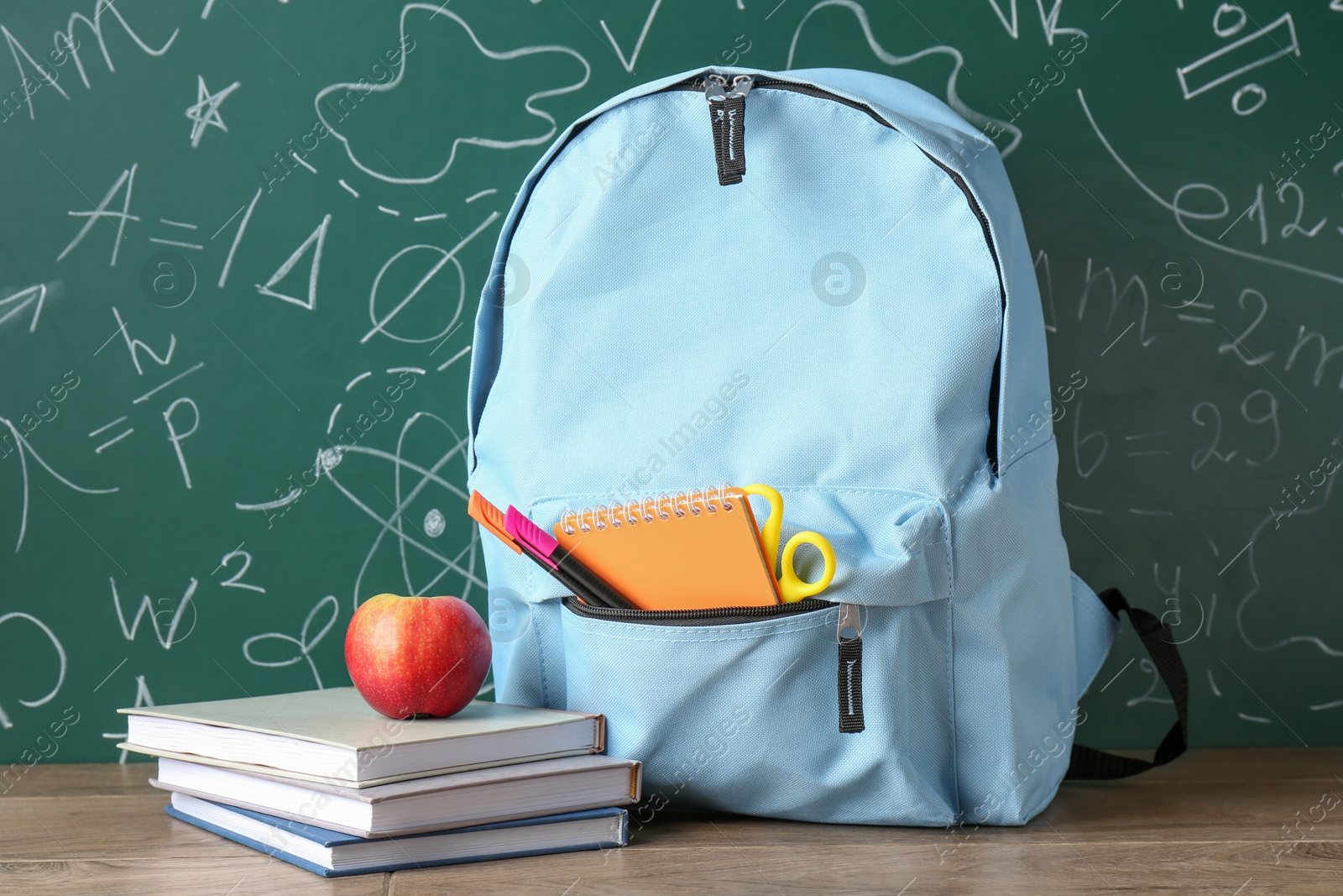 Photo of Backpack with different school stationery and apple on wooden table against green chalkboard