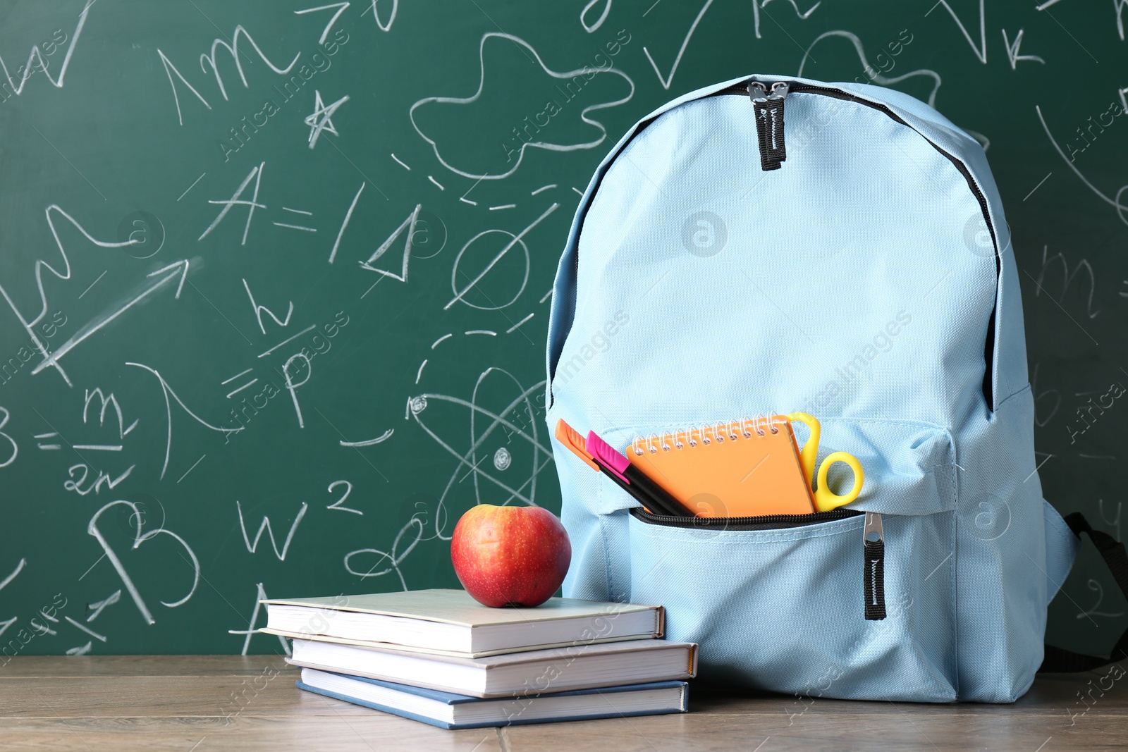 Photo of Backpack with different school stationery and apple on wooden table against green chalkboard