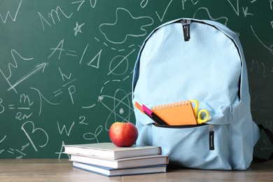 Photo of Backpack with different school stationery and apple on wooden table against green chalkboard