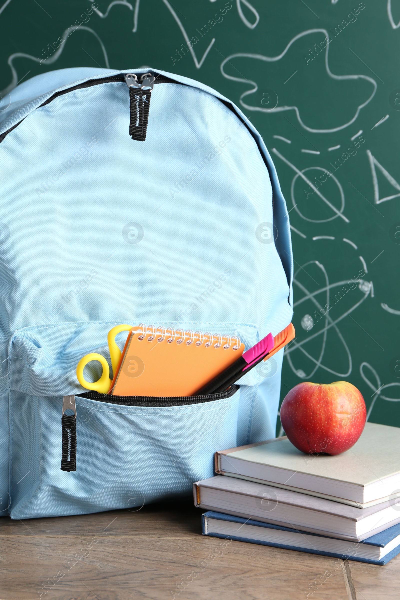 Photo of Backpack with different school stationery and apple on wooden table against green chalkboard