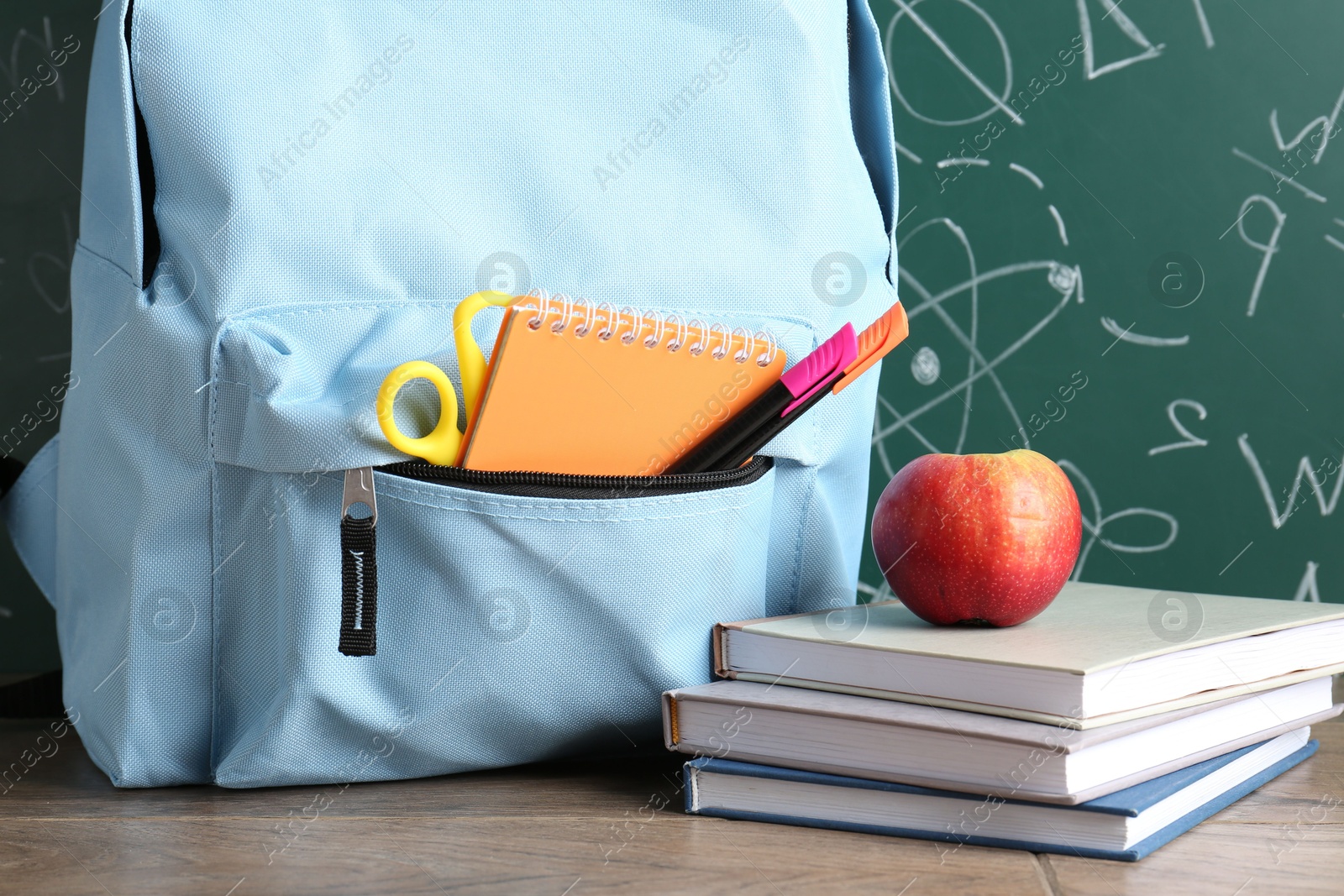 Photo of Backpack with different school stationery and apple on wooden table against green chalkboard