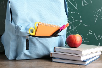 Photo of Backpack with different school stationery and apple on wooden table against green chalkboard