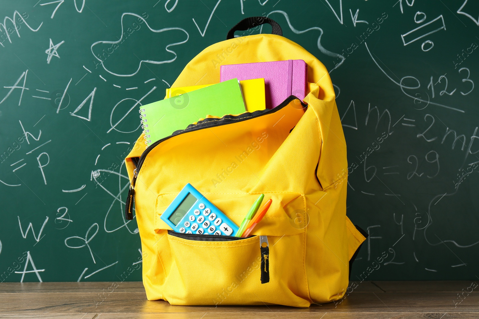 Photo of Backpack with different school stationery on wooden table against green chalkboard