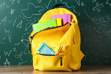 Photo of Backpack with different school stationery on wooden table against green chalkboard