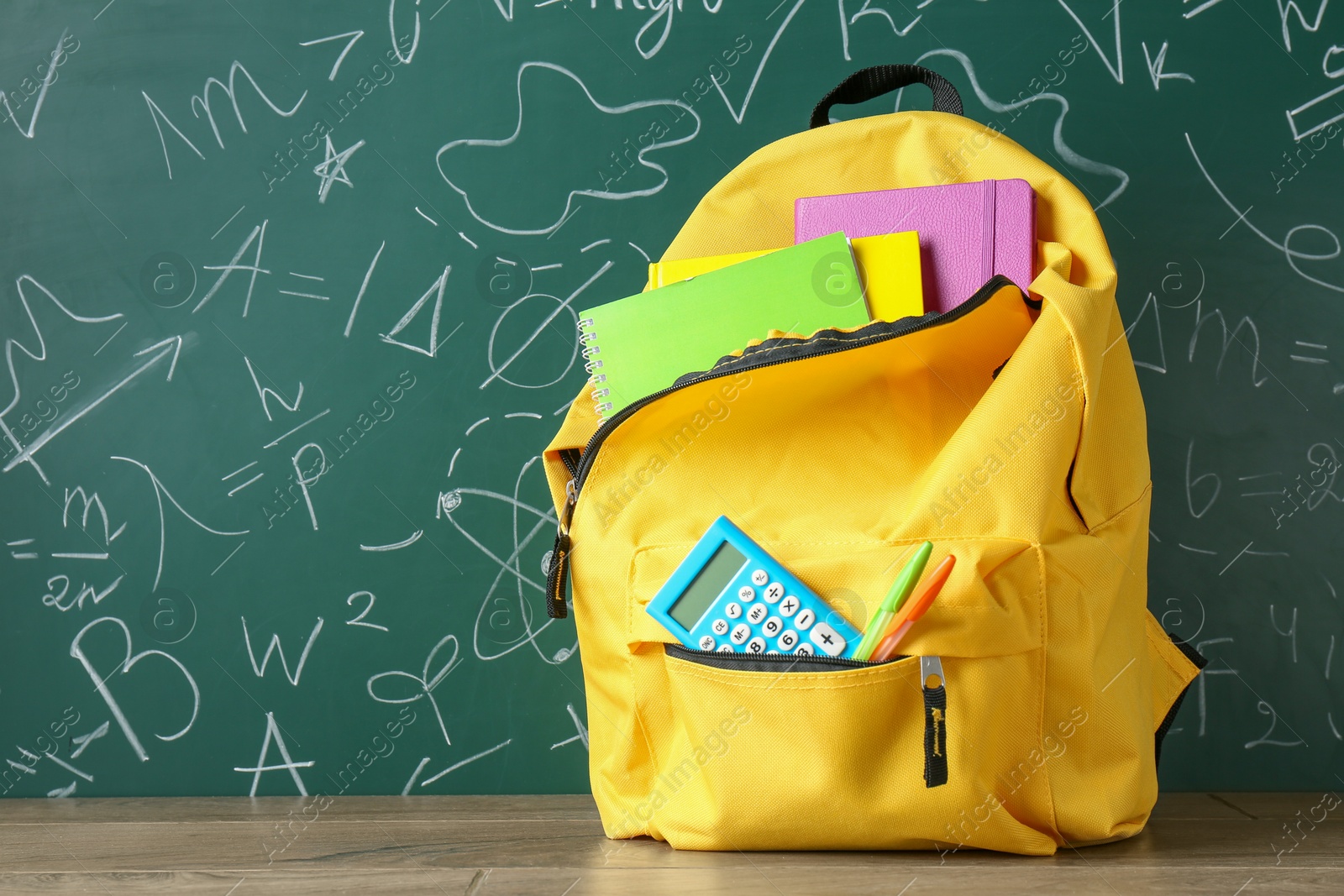 Photo of Backpack with different school stationery on wooden table against green chalkboard