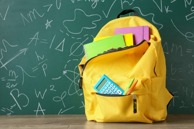 Backpack with different school stationery on wooden table against green chalkboard