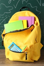 Photo of Backpack with different school stationery on wooden table against green chalkboard