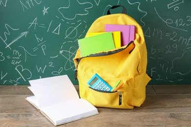 Photo of Backpack with different school stationery on wooden table against green chalkboard