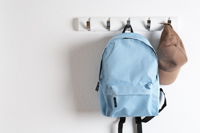 Photo of Light blue backpack and cap hanging on white wall, space for text