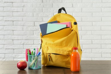 Photo of Backpack with different school stationery, apple and juice on wooden table against brick wall