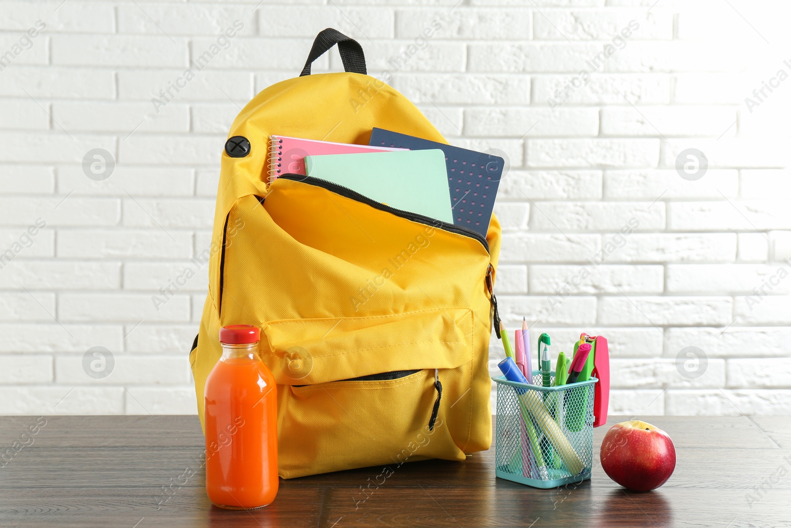 Photo of Backpack with different school stationery, apple and juice on wooden table against brick wall