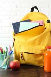 Photo of Backpack with different school stationery, apple and juice on wooden table against brick wall