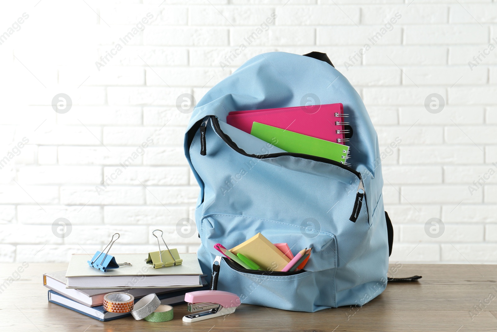 Photo of Backpack with different school stationery on wooden table against brick wall