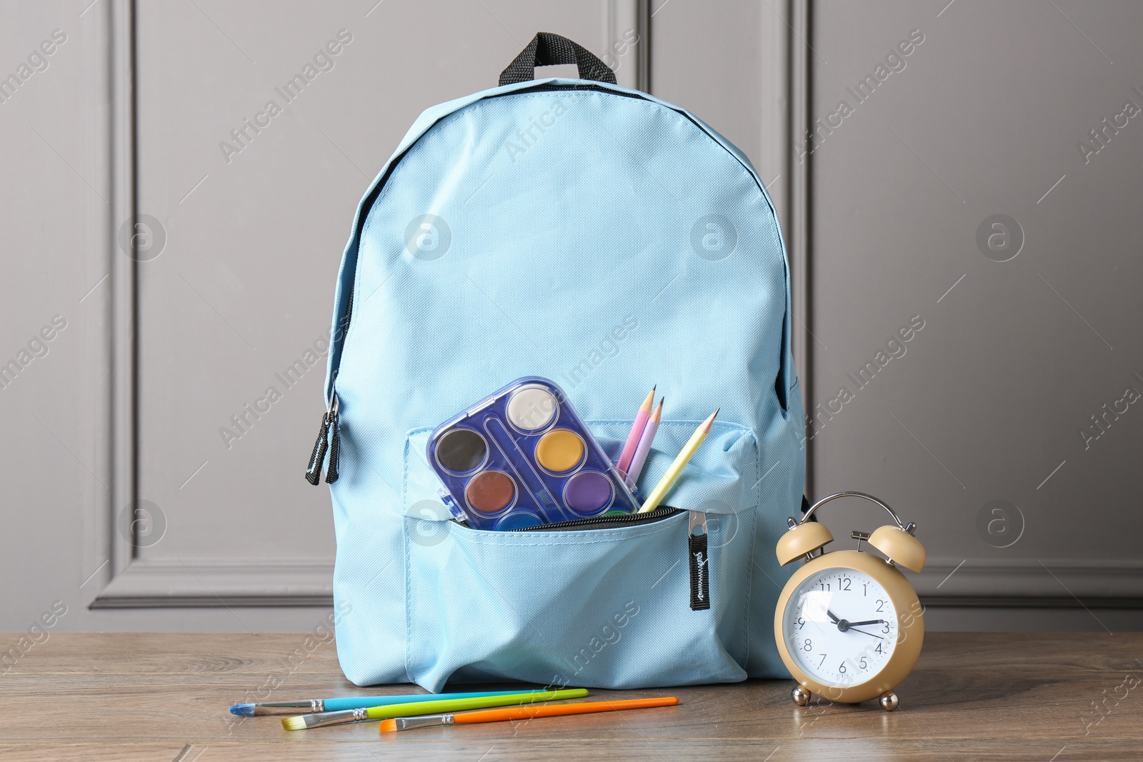 Photo of Backpack with different school stationery and alarm clock on wooden table near grey wall