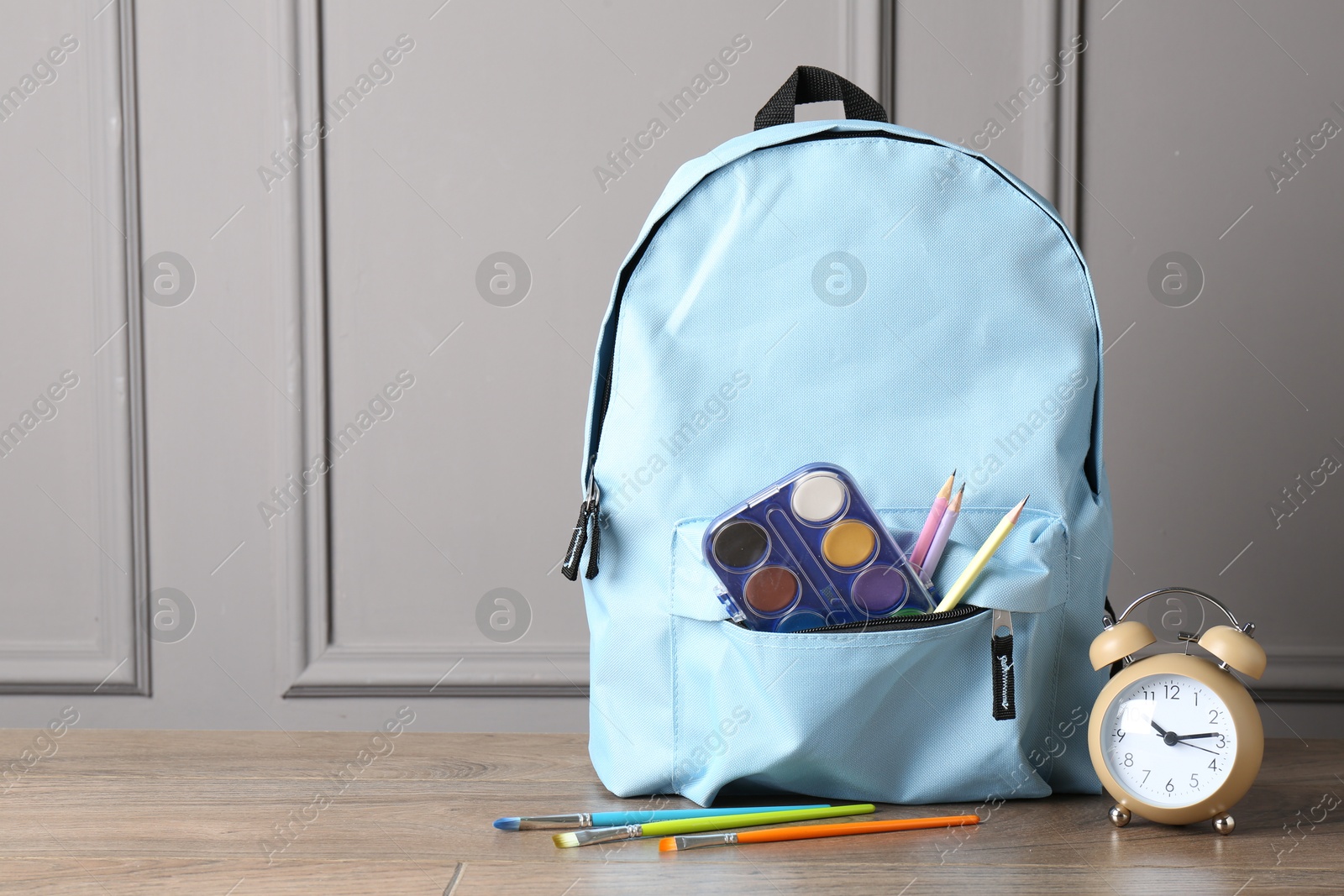 Photo of Backpack with different school stationery and alarm clock on wooden table near grey wall, space for text