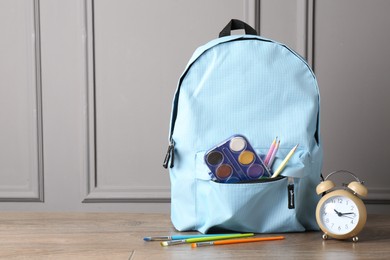 Photo of Backpack with different school stationery and alarm clock on wooden table near grey wall, space for text