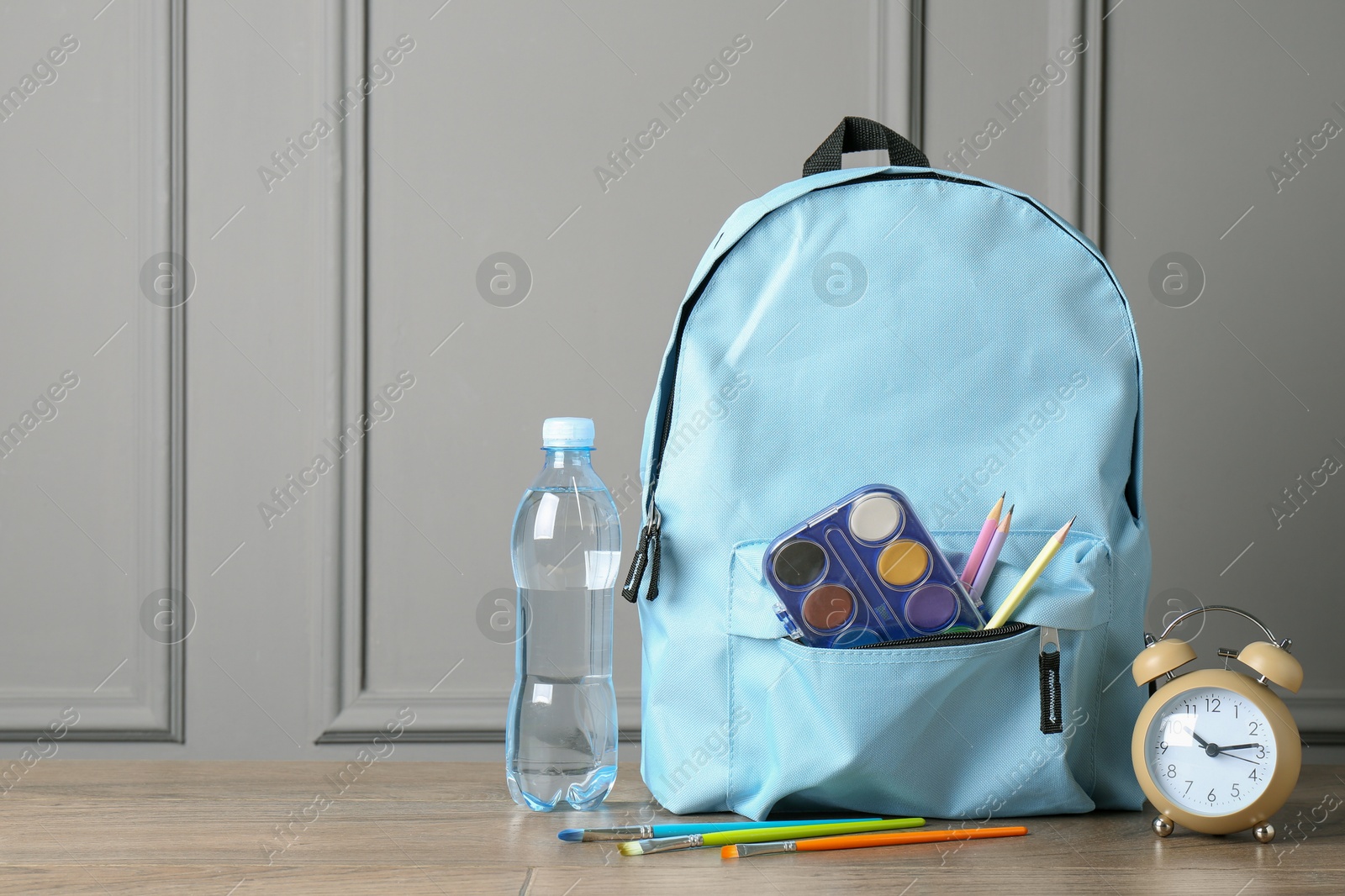 Photo of Backpack with different school stationery, bottle of water and alarm clock on wooden table near grey wall