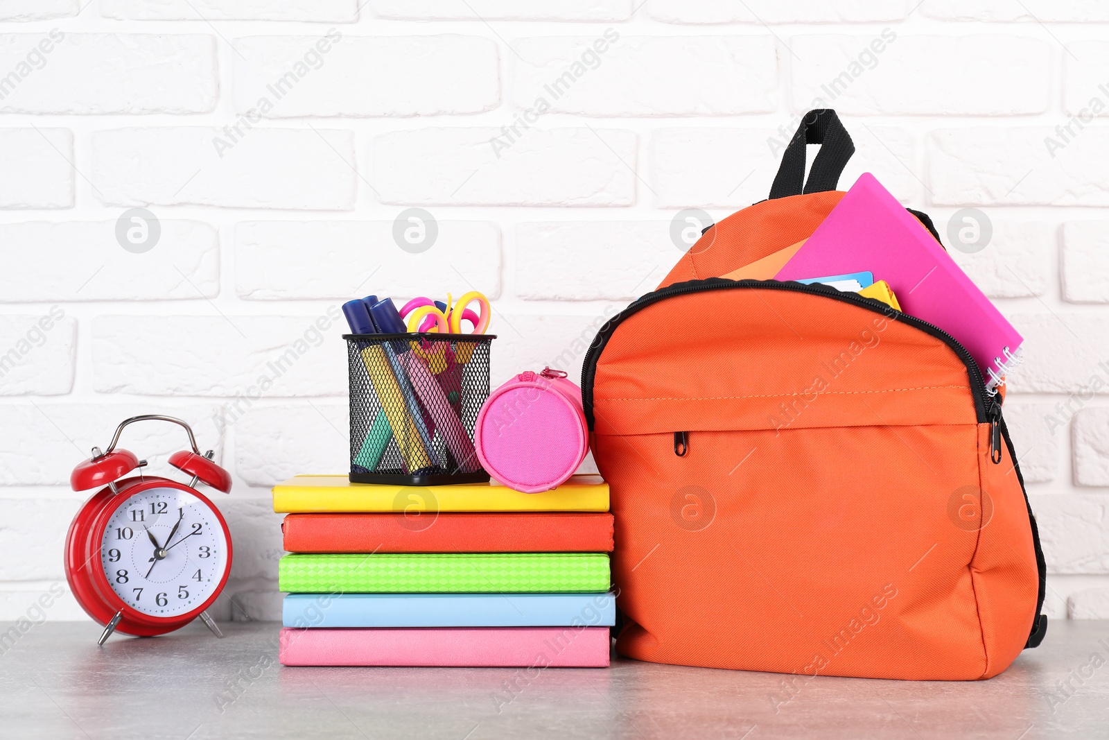 Photo of Backpack with different school stationery and alarm clock on light grey table near brick wall