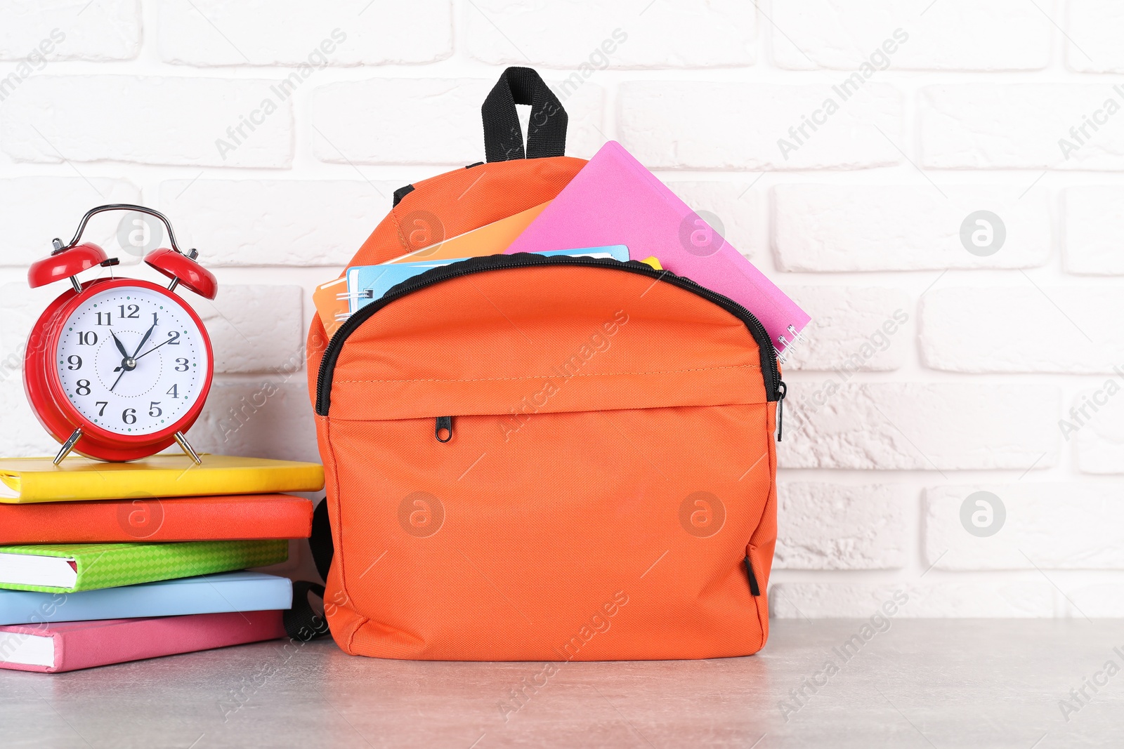 Photo of Backpack with different school stationery and alarm clock on light grey table near brick wall. Space for text