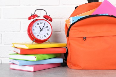 Photo of Backpack with different school stationery and alarm clock on light grey table near brick wall, closeup