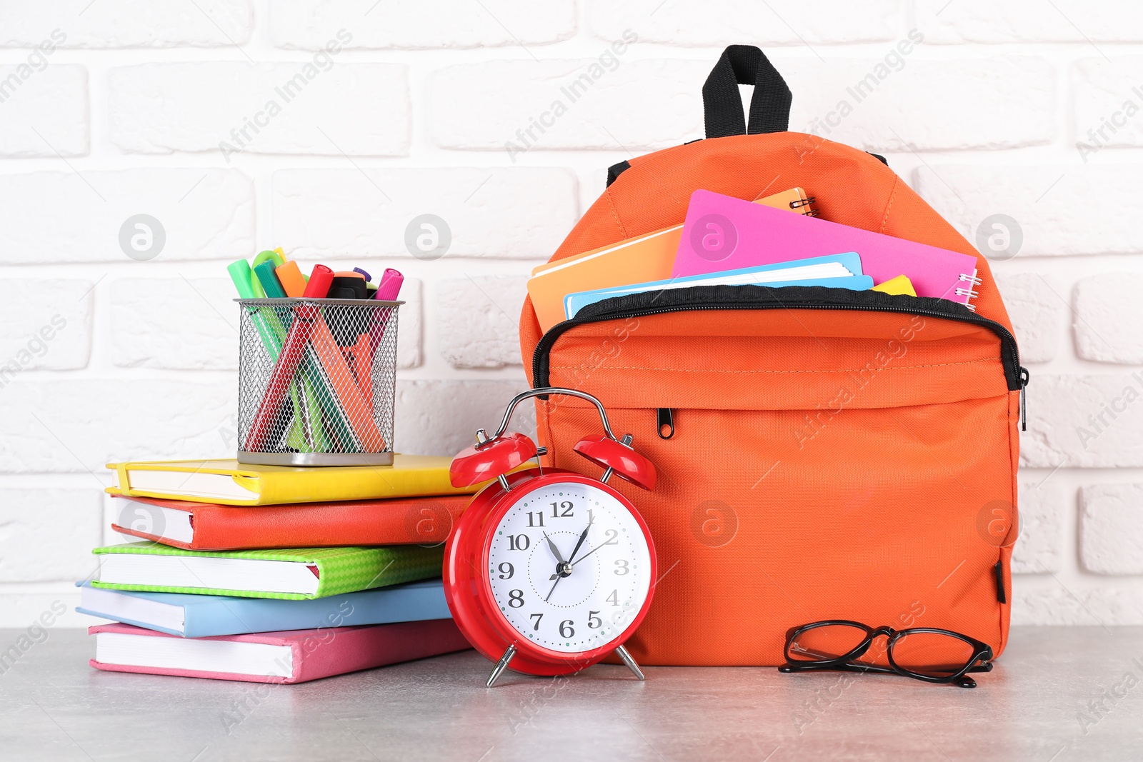 Photo of Backpack with different school stationery and alarm clock on light grey table near brick wall