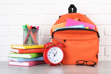 Photo of Backpack with different school stationery and alarm clock on light grey table near brick wall