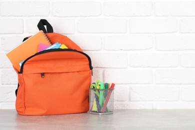 Photo of Backpack with different school stationery on light grey table near brick wall. Space for text