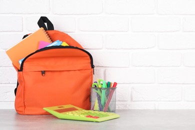 Backpack with different school stationery on light grey table near brick wall. Space for text