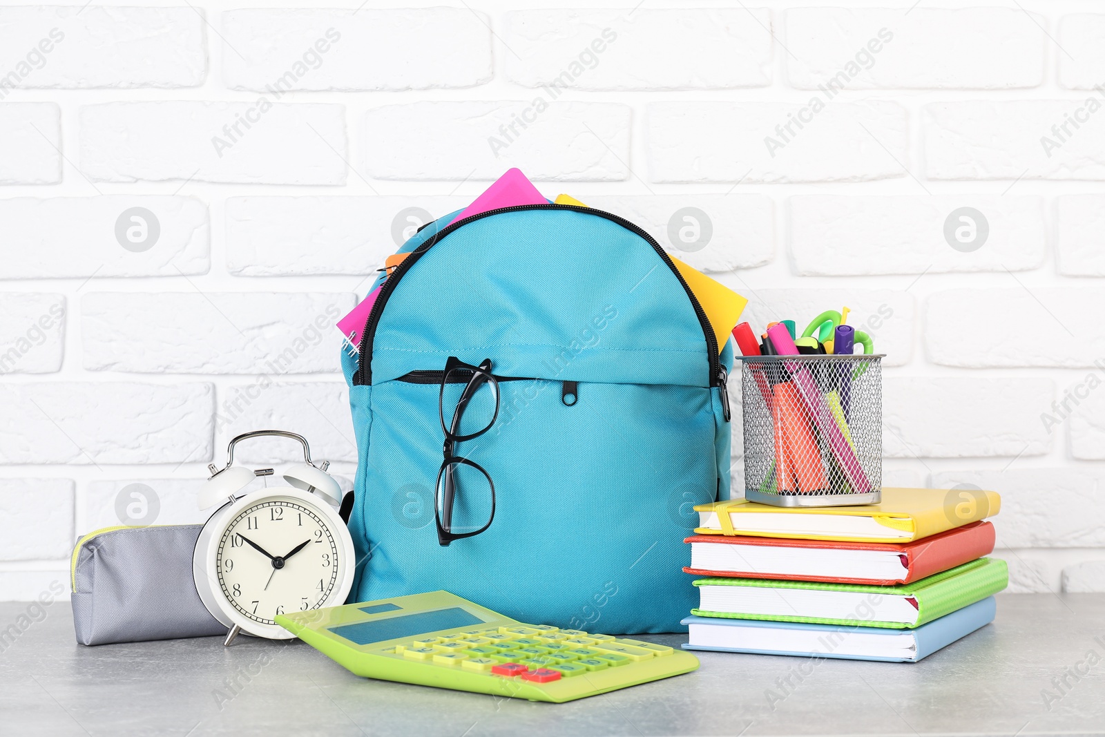 Photo of Backpack with different school stationery and alarm clock on light grey table near brick wall