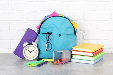 Photo of Backpack with different school stationery and alarm clock on light grey table near brick wall