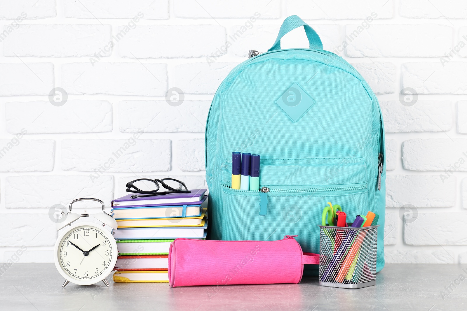 Photo of Backpack with different school stationery and alarm clock on light grey table near brick wall