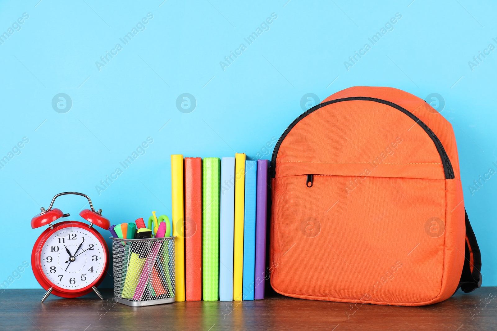 Photo of Backpack with different school stationery and alarm clock on wooden table against light blue background