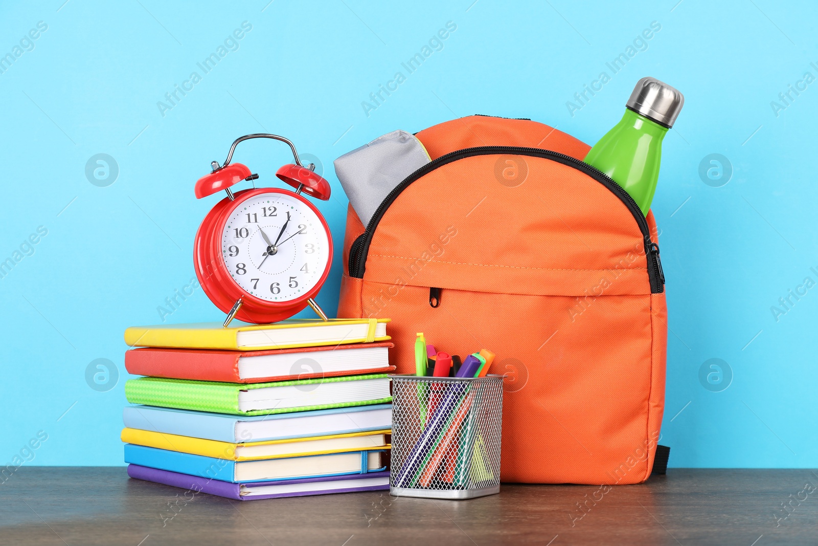 Photo of Backpack with different school stationery and alarm clock on wooden table against light blue background