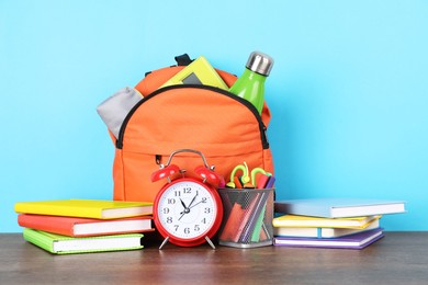 Photo of Backpack with different school stationery and alarm clock on wooden table against light blue background