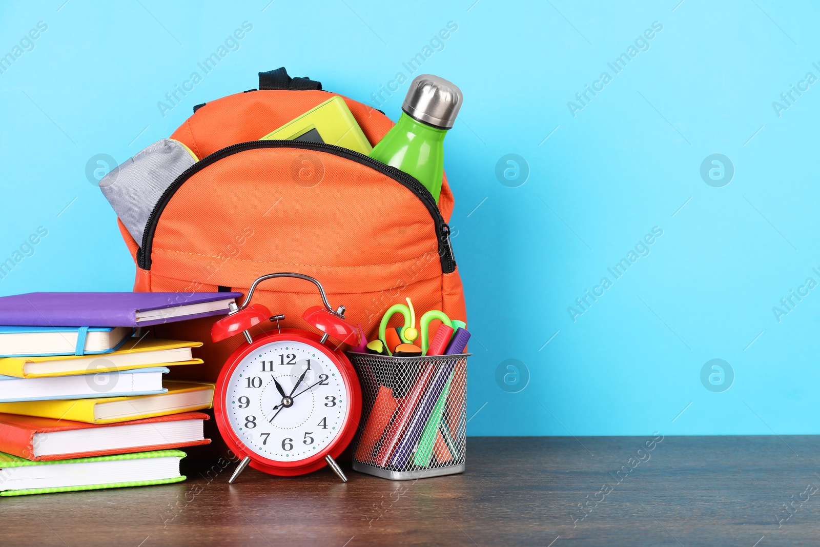 Photo of Backpack with different school stationery and alarm clock on wooden table against light blue background. Space for text