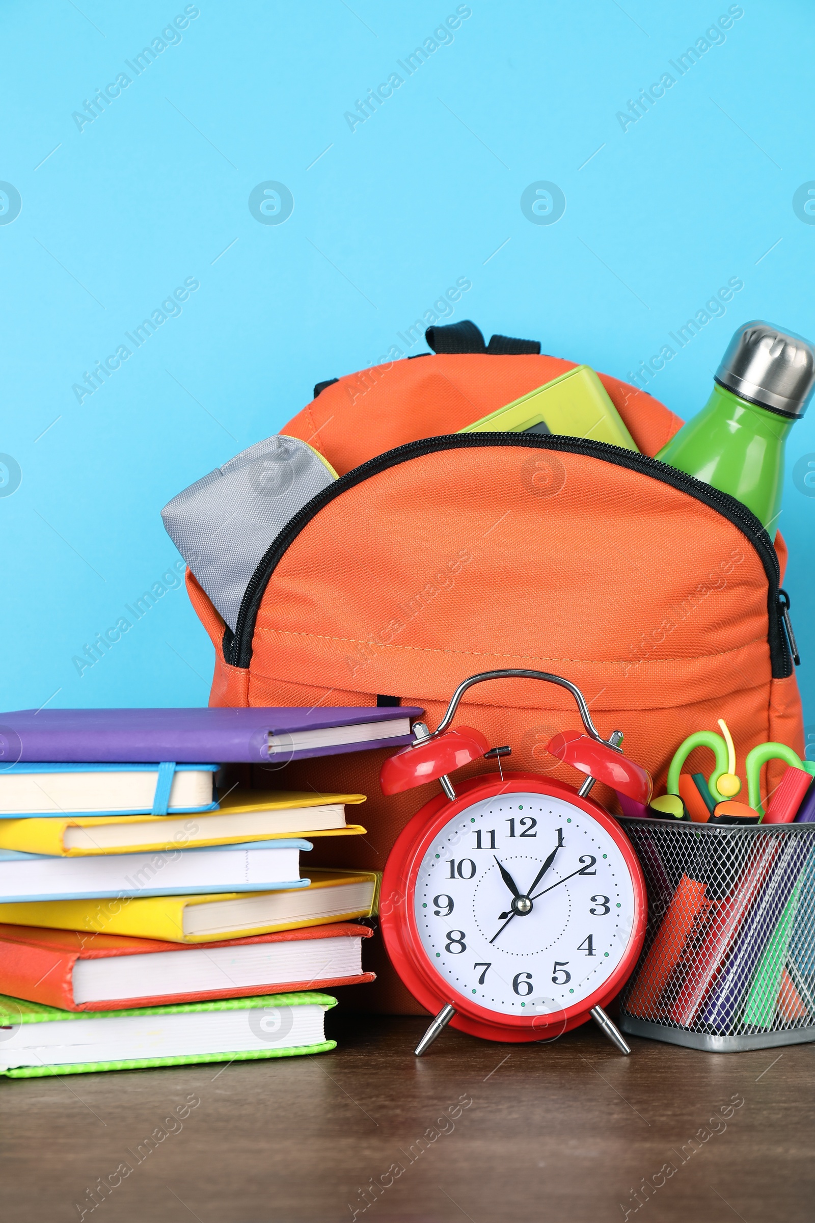 Photo of Backpack with different school stationery and alarm clock on wooden table against light blue background