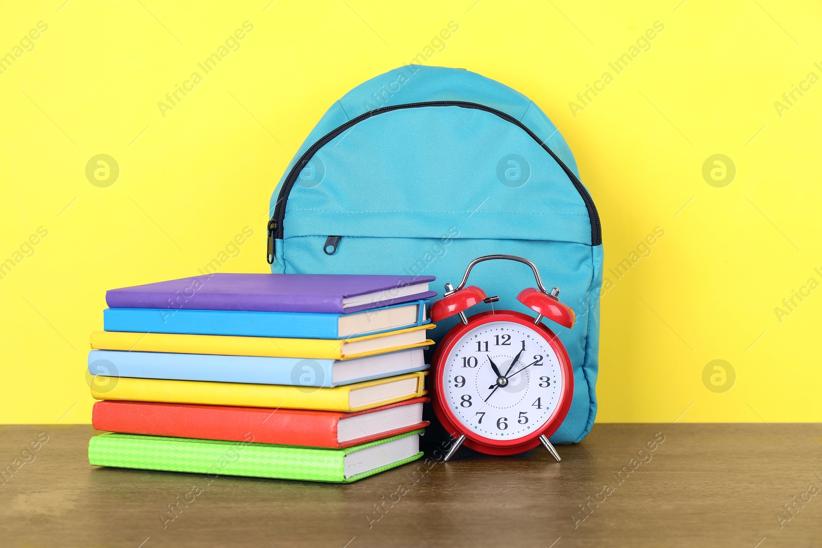 Photo of Backpack with different school stationery and alarm clock on wooden table against yellow background