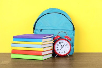 Photo of Backpack with different school stationery and alarm clock on wooden table against yellow background