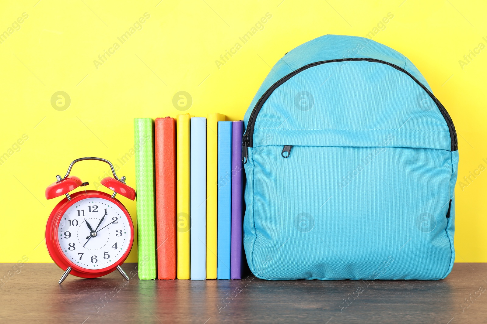 Photo of Backpack with different school stationery and alarm clock on wooden table against yellow background