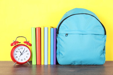 Photo of Backpack with different school stationery and alarm clock on wooden table against yellow background