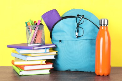 Photo of Backpack with different school stationery and bottle on wooden table against yellow background