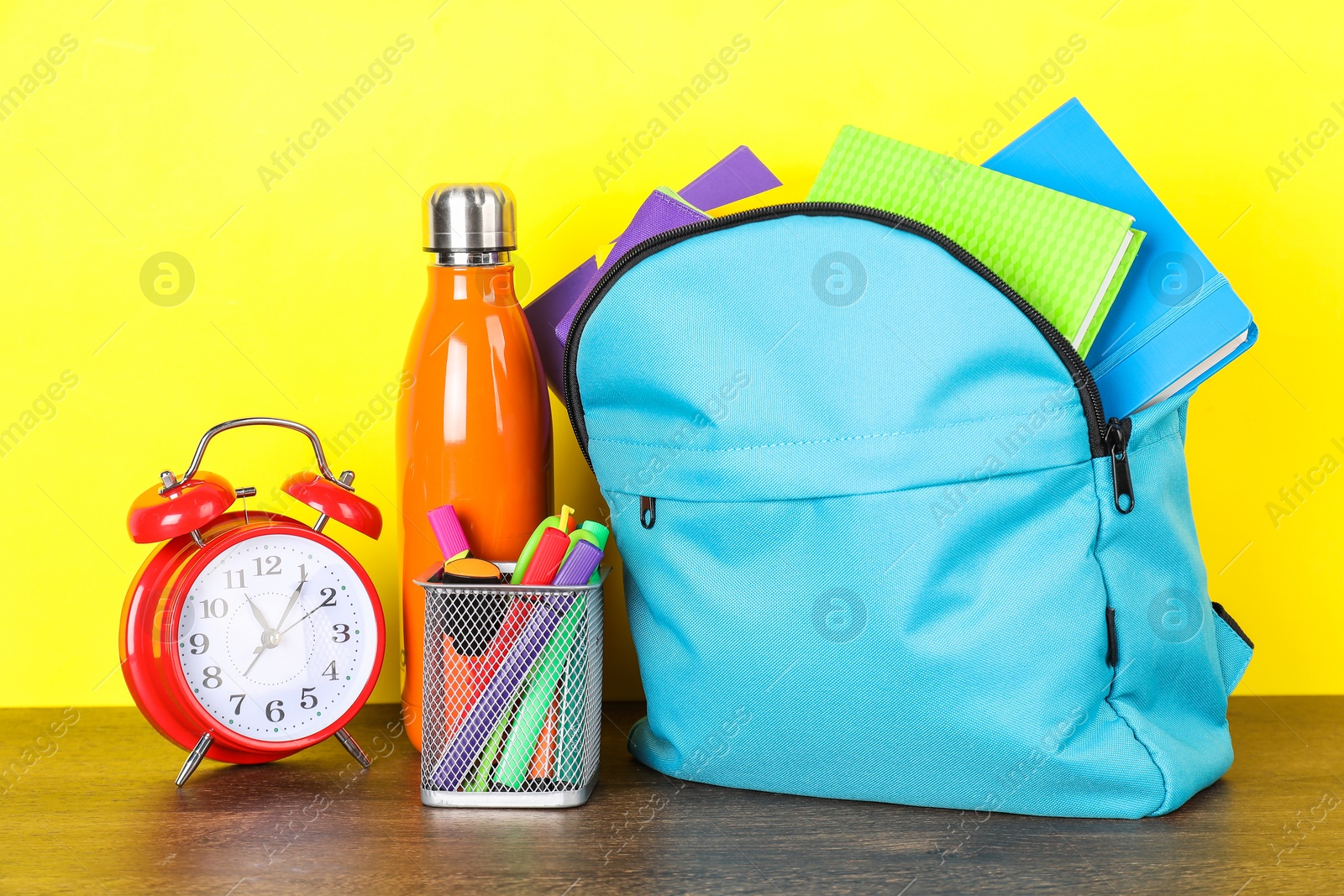 Photo of Backpack with different school stationery, bottle and alarm clock on wooden table against yellow background